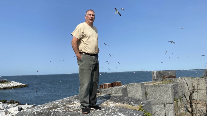 A person wearing a t-shirt tucked into jeans stand on a rock by the water