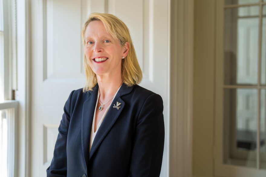 A woman wearing a navy blue blazer with a University of Virginia pin affixed to the lapel is seen in the photograph. She is smiling.