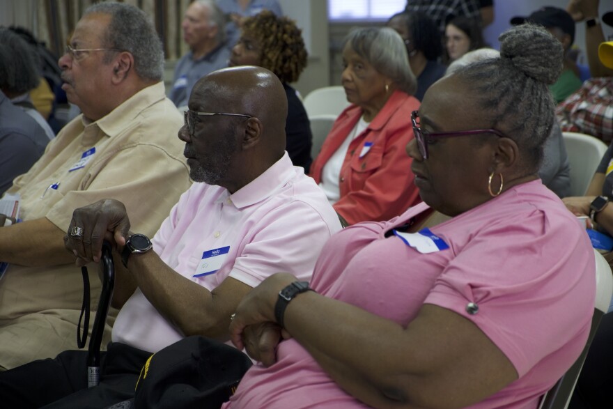 An elderly black couple listens to a presentation. A bald husband with a cane and a wife with her hair tied up in a bun, both wearing glasses and pink shirts.