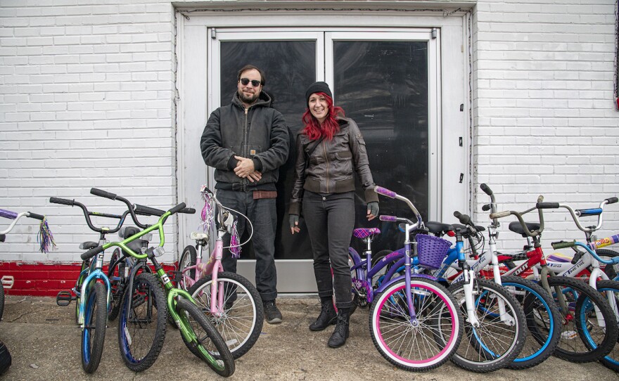 Two people wearing winter coats stand outside a building flanked by bicycles.
