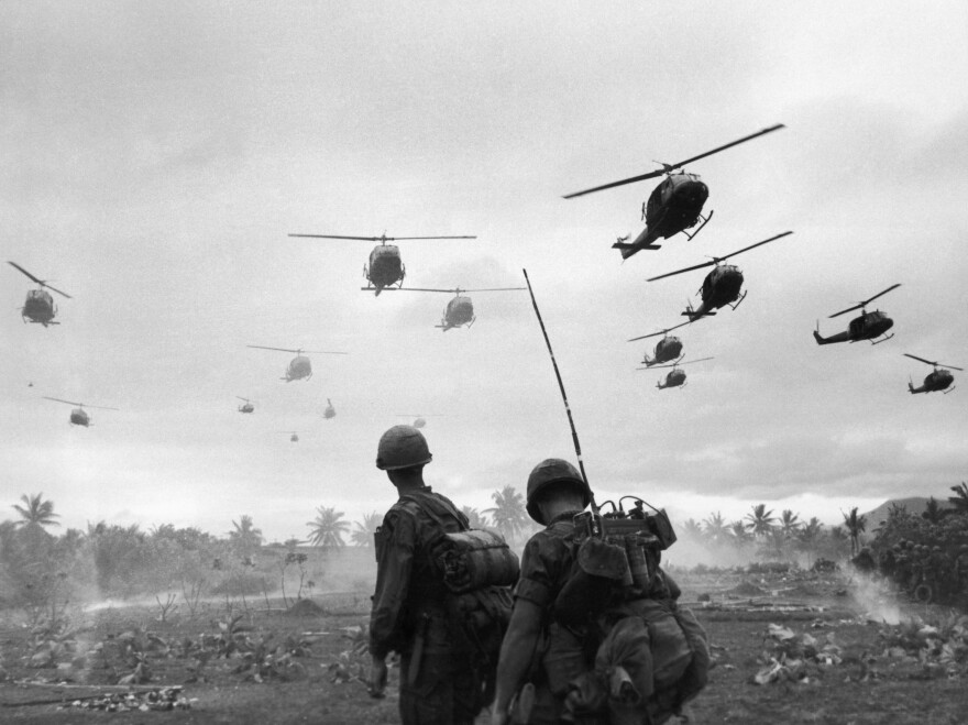 The second wave of combat helicopters of the 1st Air Cavalry Division fly over an RTO and his commander on an isolated landing zone during Operation Pershing, a search and destroy mission on the Bong Son Plain and An Lao Valley of South Vietnam, during the Vietnam War. The two American soldiers are waiting for the second wave to come in.