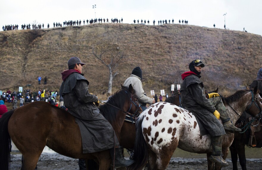 Protesters gather at Standing Rock Reservation on Thanksgiving Day to build a bridge to Turtle Island, which they consider sacred ground. Police are seen lining the island hill beyond them.