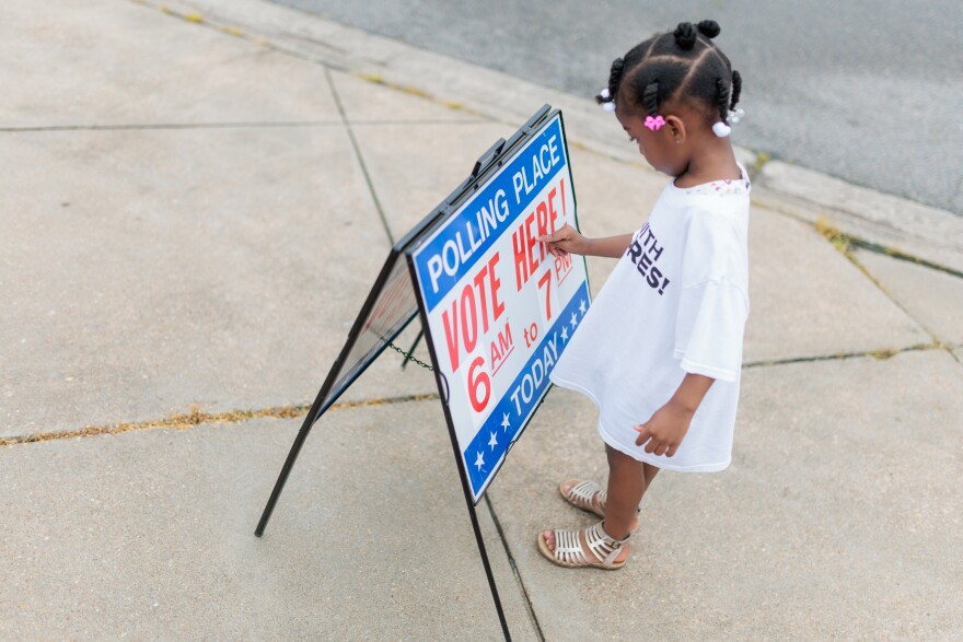 A small child is seen playing with a sign that reads "Polling Place Vote Here 6 am to 7 pm TODAY"