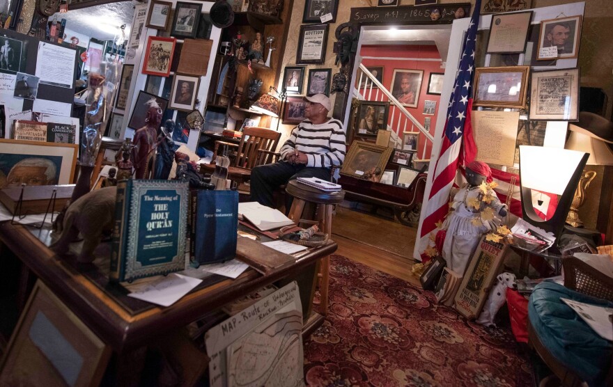 A person wearing a striped sweater sits in a room crowded with historic object. In the foreground are two books titled "THE HOLY QURAN" and "The Nyew Testament"
