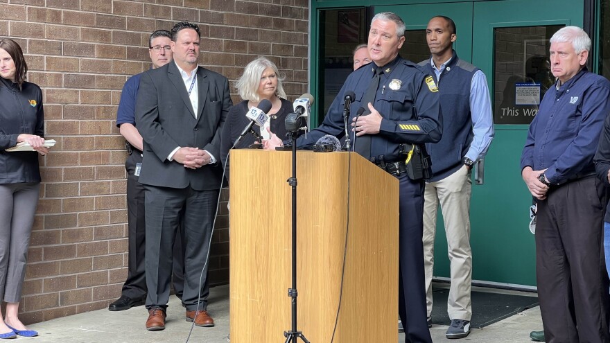 A group of people gathered at a doorway. One in a police uniform stand at a podium and speaks into a microphone