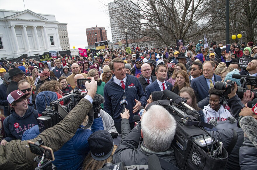 A person stand in the middle of cameras and microphones with a crowd behind.
