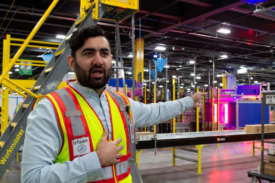 A person wearing a high-visibility vest points to a machine in a warehouse. Their vest says "Irfaan"