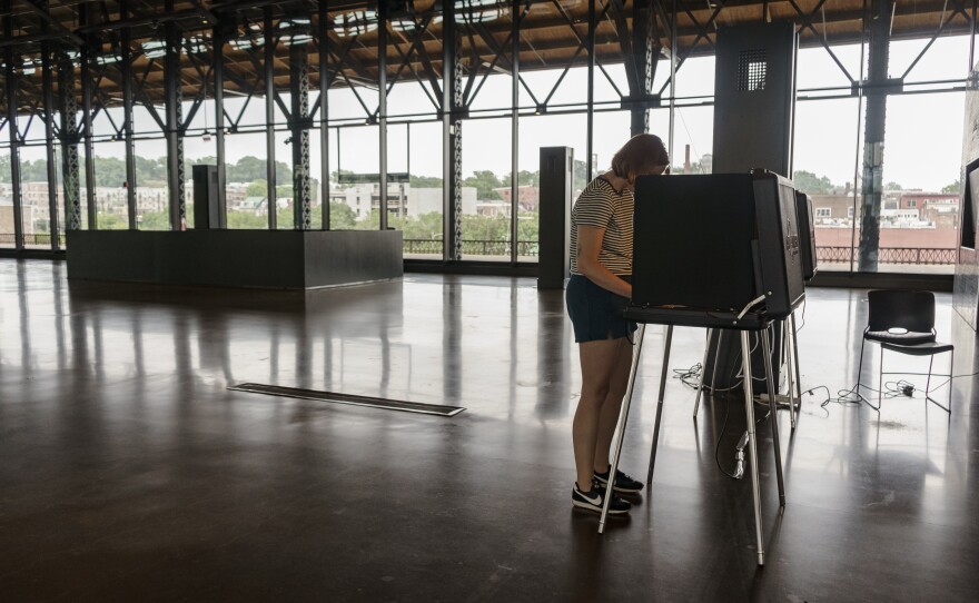 A person wearing a striped shirt stands at a voting booth in a room with cement floors and large windows